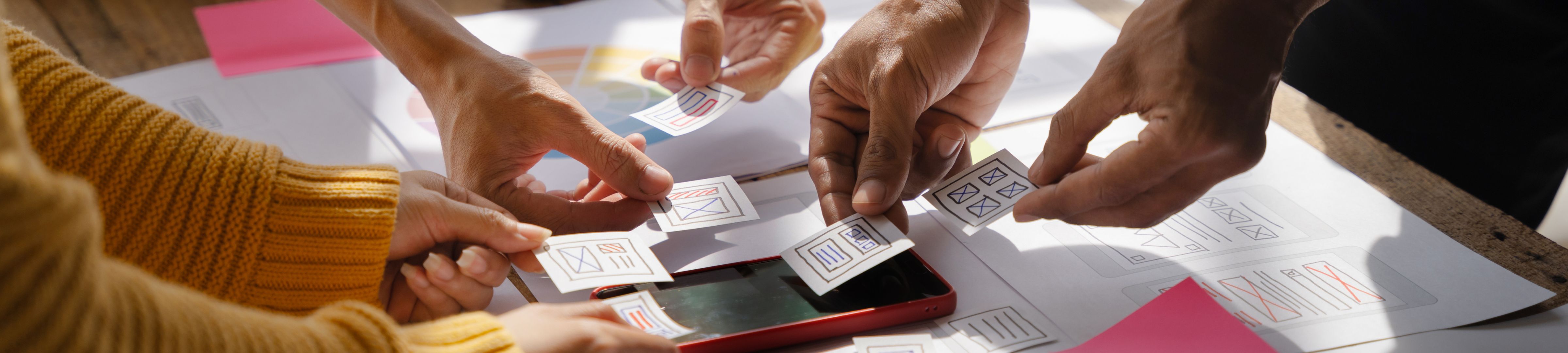 Multiple hands are seen arranging small paper cutouts with wireframe-style sketches on a table. 
