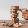 a hand carefully pulling a wooden block from a stacked Jenga tower. The tower is made of wooden blocks arranged in alternating layers on a wooden surface, with some fallen blocks scattered around. 
