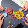 Several hands working together to assemble colourful interlocking gears on a dark gray surface