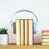 A pair of grey over-ear headphones placed around five upright hardcover books, resembling the concept of audiobooks. The books have colourful spines with yellow, green, blue, orange, and purple accents. 