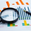 A report displaying bar and line graphs in various colours, including blue, orange, green, and yellow. A magnifying glass is placed over part of the graphs, emphasizing data analysis. 